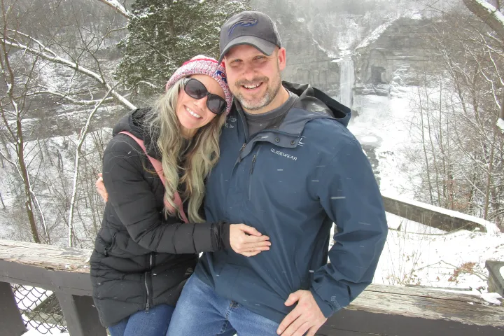 Smiling couple in winter clothing at a snowy viewpoint near a waterfall.